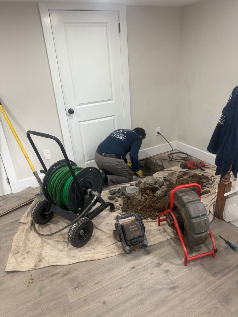 A drain machine and hose reel set up on protective sheeting inside a house, beside an opened section of floor.