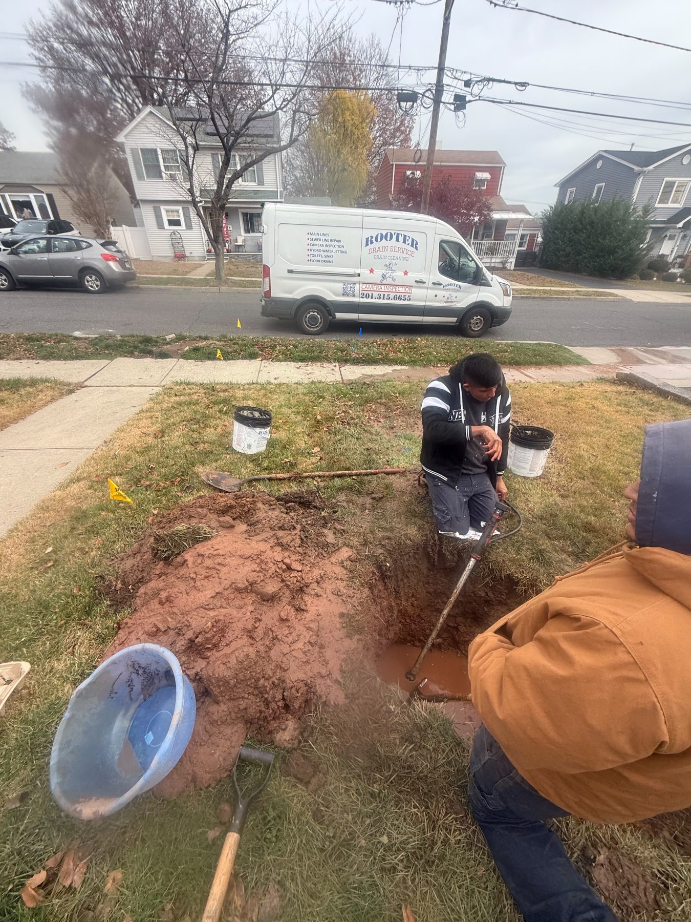 Two technicians working in an excavation in a front lawn, with buckets and a shovel alongside and the van at the kerb.